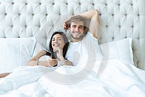 Happy couple with remote lying in bed at home and watching tv.