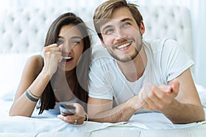 Happy couple with remote lying in bed at home and watching tv.