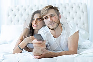 Happy couple with remote lying in bed at home and watching tv.