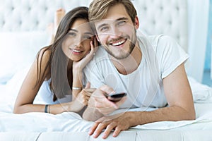 Happy couple with remote lying in bed at home and watching tv.