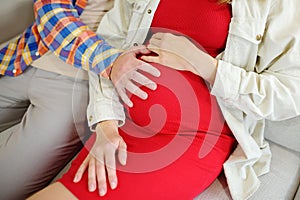 Happy couple is resting on the sofa at home. Beautiful pregnant woman and her husband touching the belly and feeling the movements