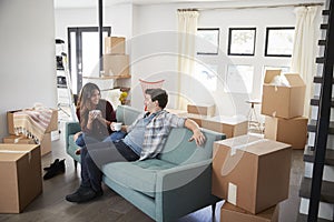 Happy Couple Resting On Sofa Surrounded By Boxes In New Home On Moving Day