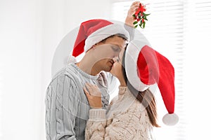 Happy couple in Santa hats kissing under mistletoe bunch at home