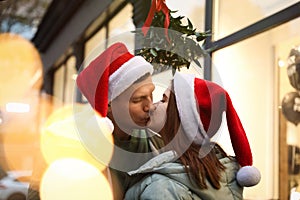 Happy couple in Santa hats kissing under mistletoe bunch outdoors, bokeh effect