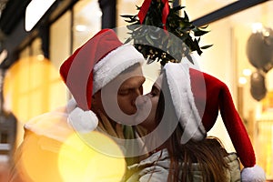 Happy couple in Santa hats kissing under mistletoe bunch outdoors, bokeh effect