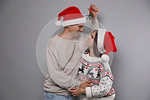 Happy couple in Santa hats standing under mistletoe bunch on grey background