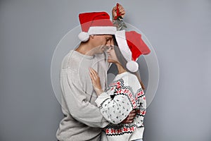 Happy couple in Santa hats standing under mistletoe bunch on grey background