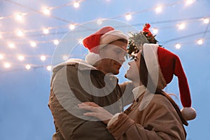 Happy couple in Santa hats standing under mistletoe bunch outdoors