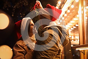 Happy couple in Santa hats standing under mistletoe bunch outdoors, bokeh effect