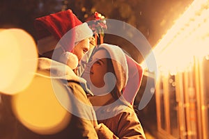 Happy couple in Santa hats standing under mistletoe bunch outdoors, bokeh effect