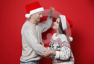 Happy couple in Santa hats standing under mistletoe bunch on red background