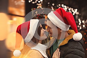 Happy couple in Santa hats standing under mistletoe wreath outdoors, bokeh effect