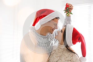 Happy couple in Santa hats under mistletoe bunch at home, bokeh effect