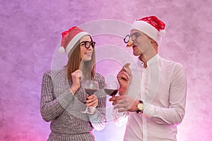 Happy couple in santa hats with wine in glasses holding mask near face and celebrating Valentine`s Day