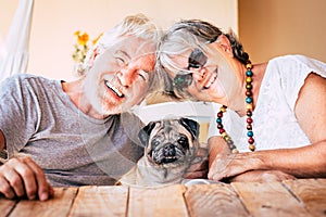 Happy couple of senior caucasian people have fun and smile with funny nice old dog pug in the middle sitting on the table together photo