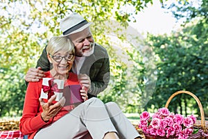 Couple senior man surprise giving gift box to his wife while relaxing in the park