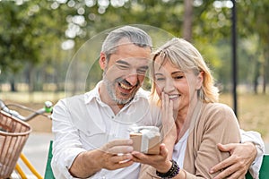 Happy couple senior man surprise giving gift box to his wife while relaxing and sitting on the bench in the park