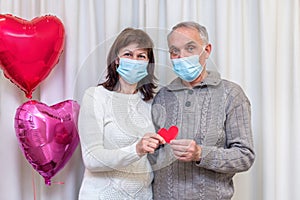 Happy couple seniors celebrate Valentine's Day in mask. Man and woman hold red heart in hands. Romantic relationships