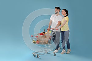 Happy couple with shopping cart full of groceries on light blue background