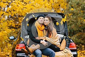 A happy couple sits on the trunk of a car, enjoying the view of autumn. The concept of travel and weekend holidays