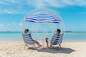 Happy couple is sitting in chairs under parasol on a tropical beach. Summer vacation concept