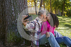 Happy couple sitting on the grass in the park kissing and taking a picture with the mobile phone