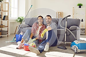 Happy couple sitting at home with vacuum cleaner and mop and looking cheerful at camera.