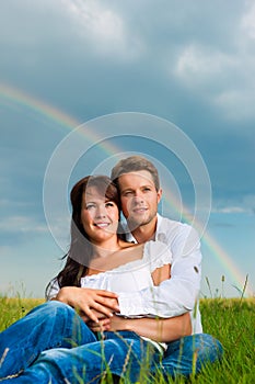 Happy couple sitting on a meadow under rainbow