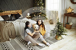 Happy couple is sitting near the bed with a dog, christmas. new year at home. family and date