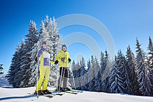 Happy couple skiers posing on skis before skiing at ski resort. Clear blue sky, snow-covered fir trees on background.