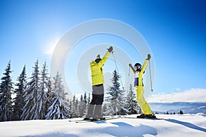 Happy couple skiers standing on mountain edge, rejoicing, raising hands up. Clear blue sky over winter mountain nature.