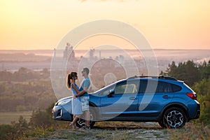 Happy couple spending time together near their SUV car during honeymoon road trip at warm summer evening. Young man and