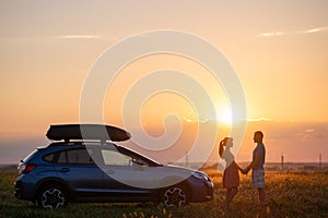 Happy couple spending time together near their SUV car during honeymoon road trip at warm summer evening. Young man and