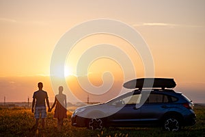 Happy couple spending time together near their SUV car during honeymoon road trip at warm summer evening. Young man and