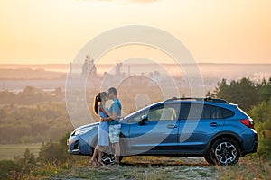 Happy couple spending time together near their SUV car during honeymoon road trip at warm summer evening. Young man and woman