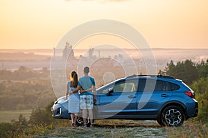 Happy couple standing near their car at sunset. Young man and woman enjoying time together travelling by vehicle