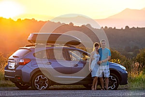 Happy couple standing near their car at sunset. Young man and woman enjoying time together travelling by vehicle