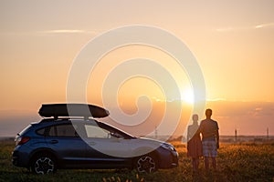 Happy couple standing near their car at sunset. Young man and woman enjoying time together travelling by vehicle