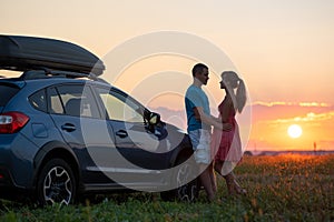 Happy couple standing near their car at sunset. Young man and woman enjoying time together travelling by vehicle