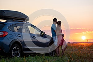 Happy couple standing near their car at sunset. Young man and woman enjoying time together travelling by vehicle