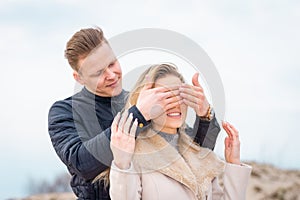 Happy couple are standing outdoor their celebrating on trip. Young man covers the smiling girl`s eyes with his hands