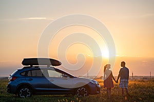 Happy couple standing beside their SUV car during honeymoon road trip at warm summer evening. Young man and woman enjoying time