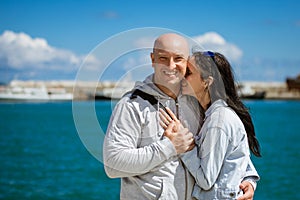 A happy couple stands on the embankment with their arms around each other against the background of the sea and sky on a Sunny day