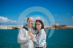 A happy couple stands on the embankment with their arms around each other against the background of the sea and sky on a Sunny day