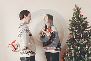 happy couple in stylish sweaters exchanging gifts in festive room at christmas tree. man holding surprise present behind back. me