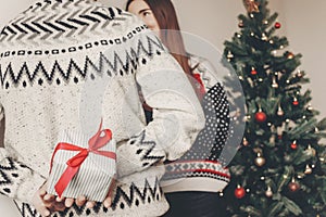 happy couple in stylish sweaters exchanging gifts in festive room at christmas tree. man holding surprise present behind back. me