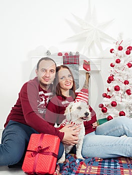 Happy couple with dog, all in Christmas clothes sitting near Christmas tree