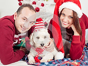 Happy couple with dog, all in Christmas clothes sitting near Christmas tree