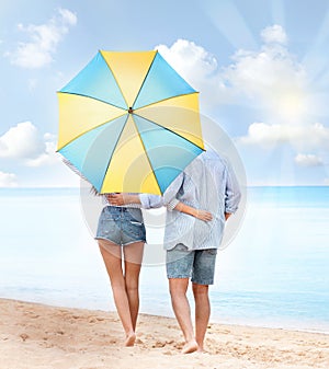 Happy couple with umbrella for sun protection walking on beach near sea, back view