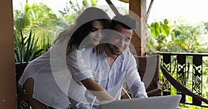 Happy couple using laptop on summer terrace typing message together, woman embracing man sitting at table happy smiling